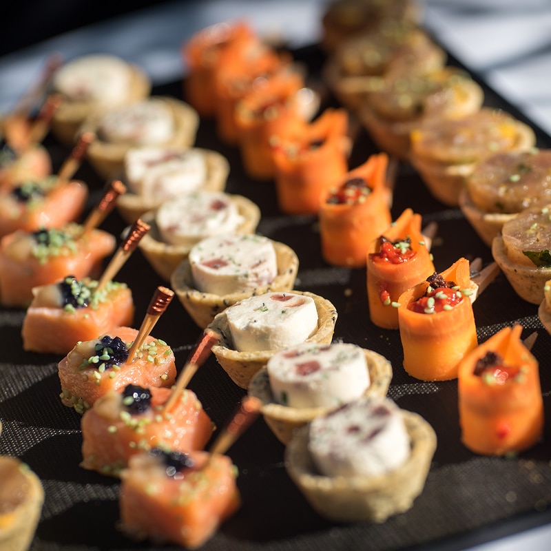 Amuse-bouches traiteur : canapés saumon, fromage, carottes Assortiment d'amuse-bouches variés présentés sur un plateau noir : saumon roulé, tartelettes au fromage frais, et rouleaux de carottes garnis.