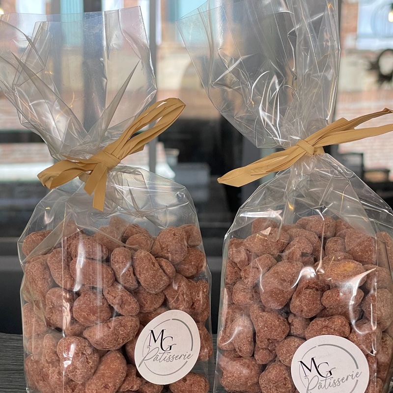 Amandes Chocolatées MG Pâtisserie Artisanales Deux sacs transparents remplis de fruits secs enrobés de chocolat ou de sucre brun, fermés par un ruban de raphia et portant un autocollant 'MG Pâtisserie'.