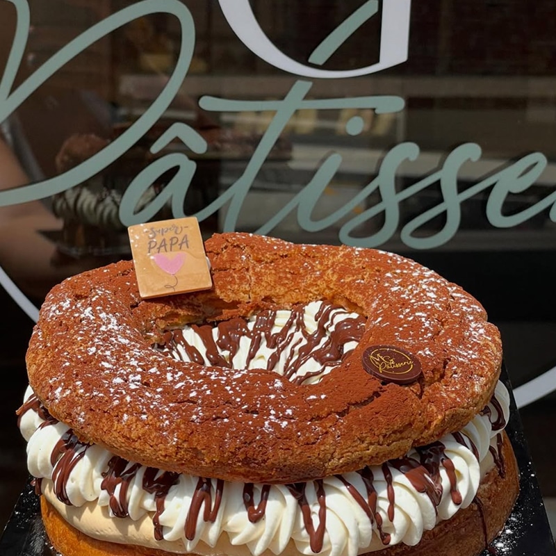 Paris-Brest Géant au Chocolat Spécial Fête des Pères Gros plan sur un Paris-Brest géant au chocolat, saupoudré de cacao et de sucre glace, avec une décoration "Super Papa" en chocolat.