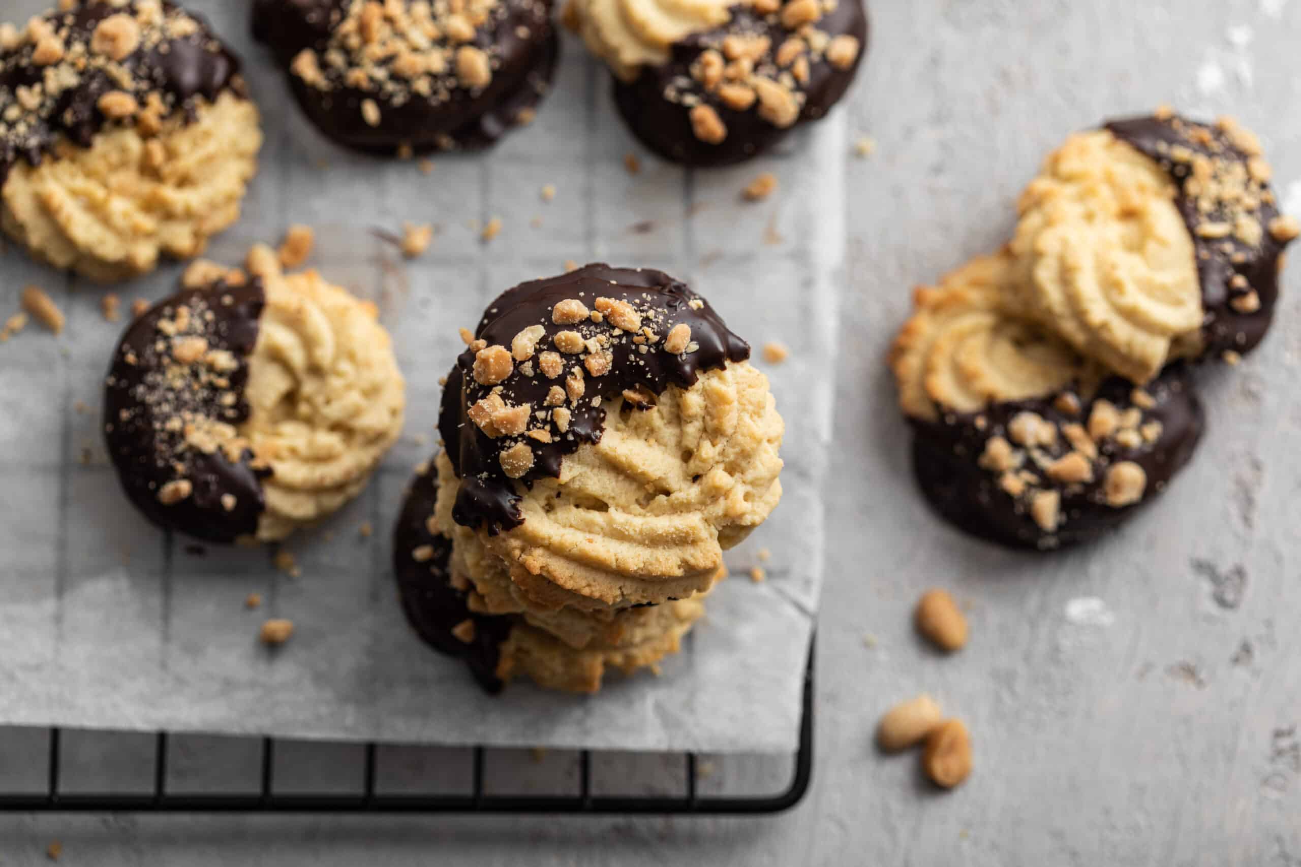 Gros plan sur un empilement de biscuits sablés en forme de rosace, trempés dans du chocolat noir et parsemés de cacahuètes concassées, posés sur une grille.