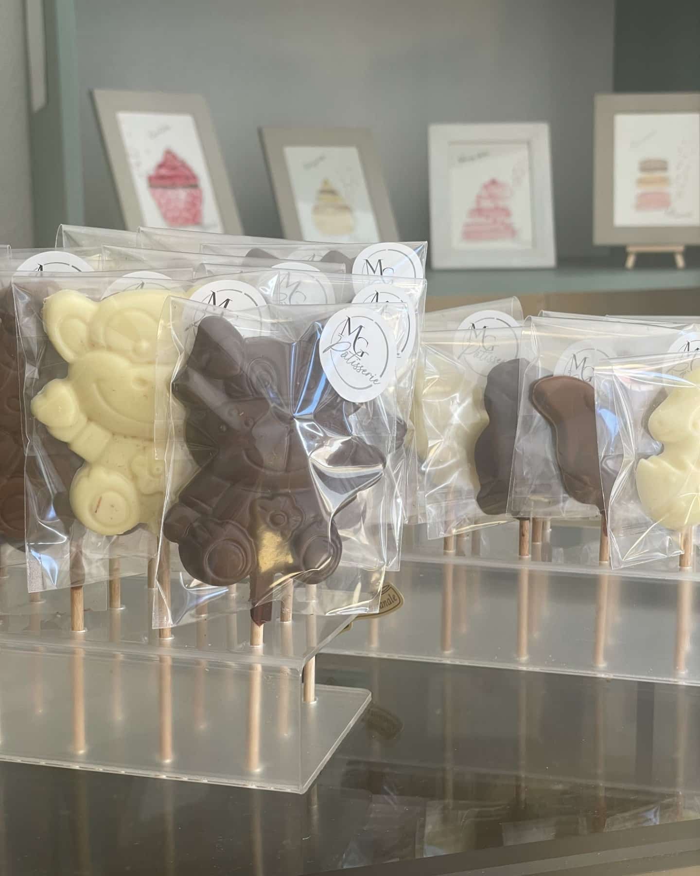 Chocolats en sucette en forme d'ours en chocolat blanc et noir, emballés individuellement avec un autocollant 'MG Pâtisserie', présentés sur un présentoir transparent.