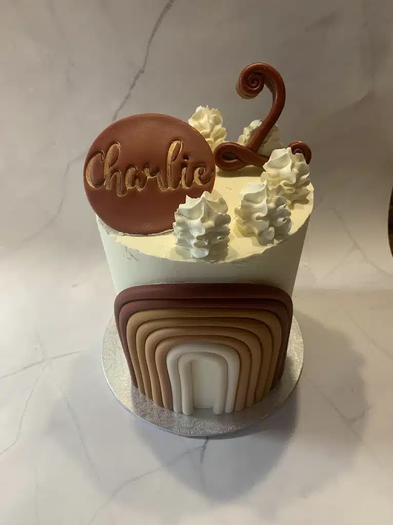 Gâteau d'anniversaire blanc décoré d'un arc-en-ciel terre cuite, d'un disque 'Charlie' marron et d'un chiffre '2' en sucre.