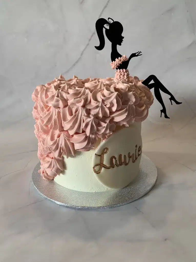 Gâteau Chic 'Laurie' : Volants Rose et Silhouette Élégante Gâteau d'anniversaire avec glaçage blanc et volants rose pâle, surmonté d'une silhouette noire de femme assise, nom 'Laurie' en or.