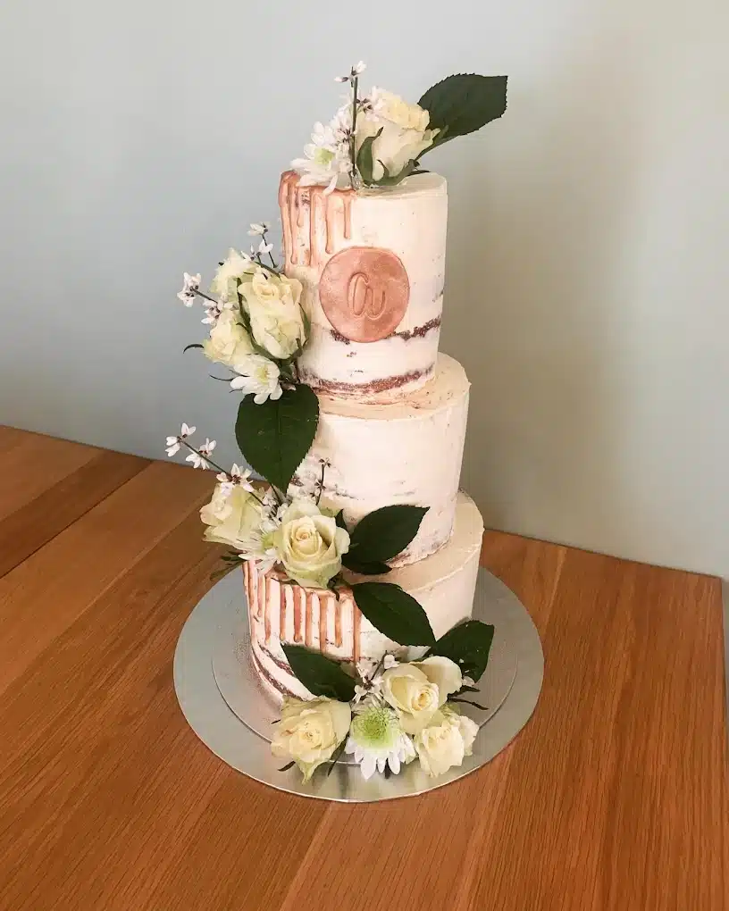 Gâteau de mariage trois étages 'naked' fleurs et or rose Gâteau de mariage à trois étages 'naked cake' avec glaçage or rose coulant, décoré de roses pâles et d'un médaillon avec la lettre 'a'.