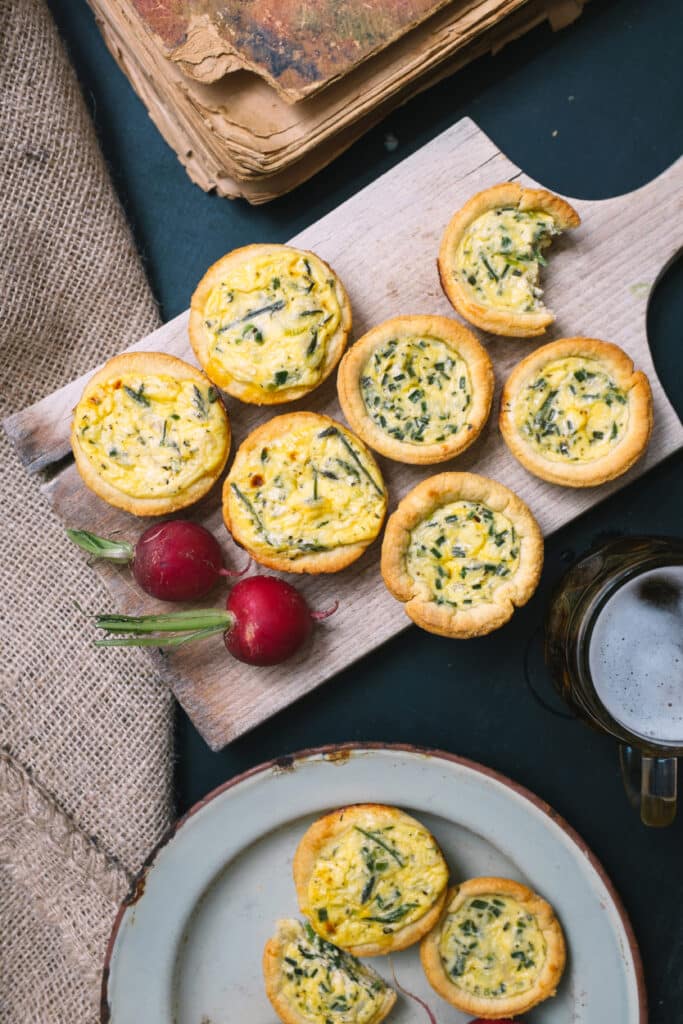 Mini Quiches Rustiques aux Œufs et Bière : Recette Fermière Mini-quiches aux œufs et ciboulette sur planche en bois rustique, près de radis, d'une bière et de vieux livres.