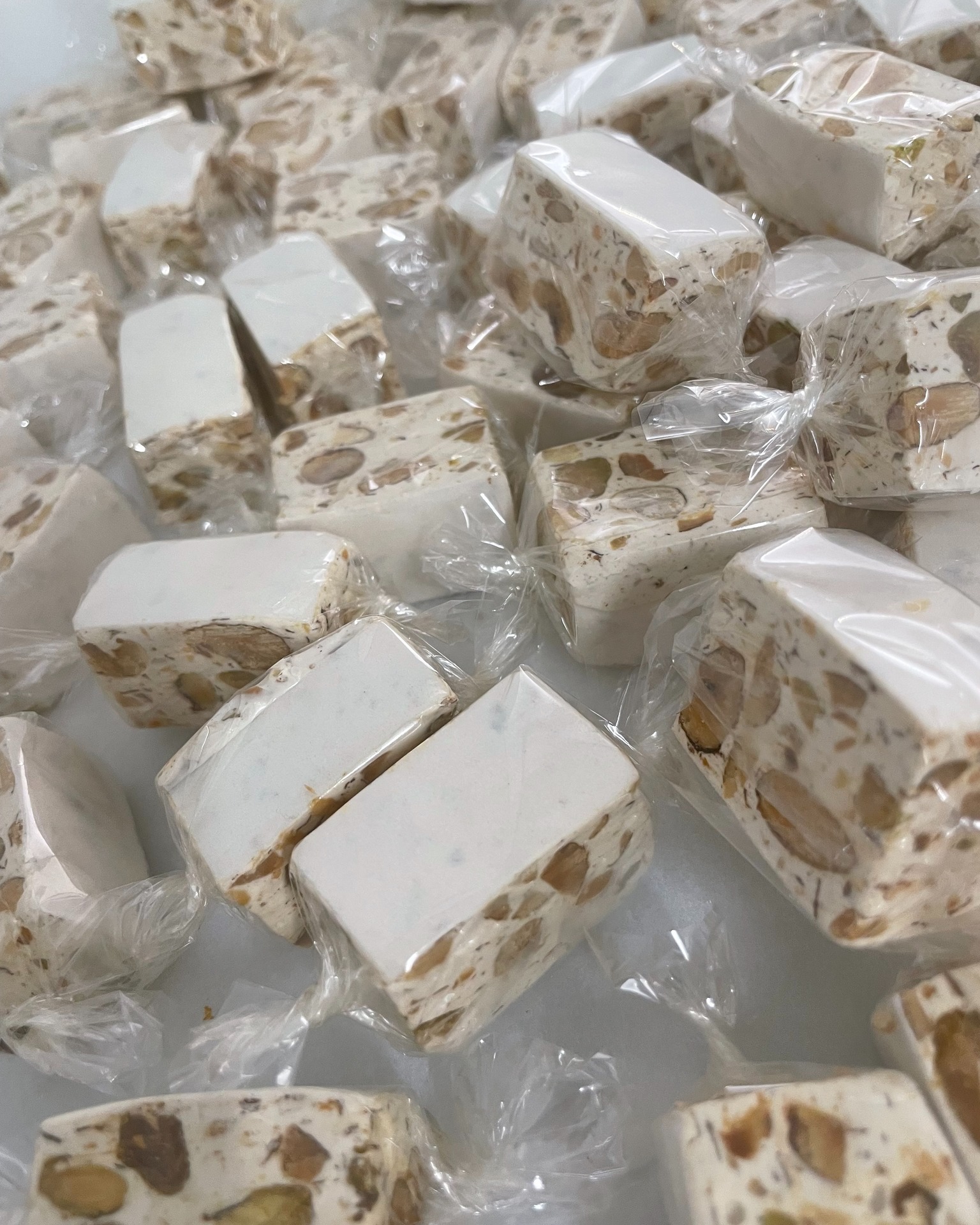 Multiples morceaux de nougat blanc artisanal aux amandes et pistaches, emballés individuellement dans du film plastique transparent.