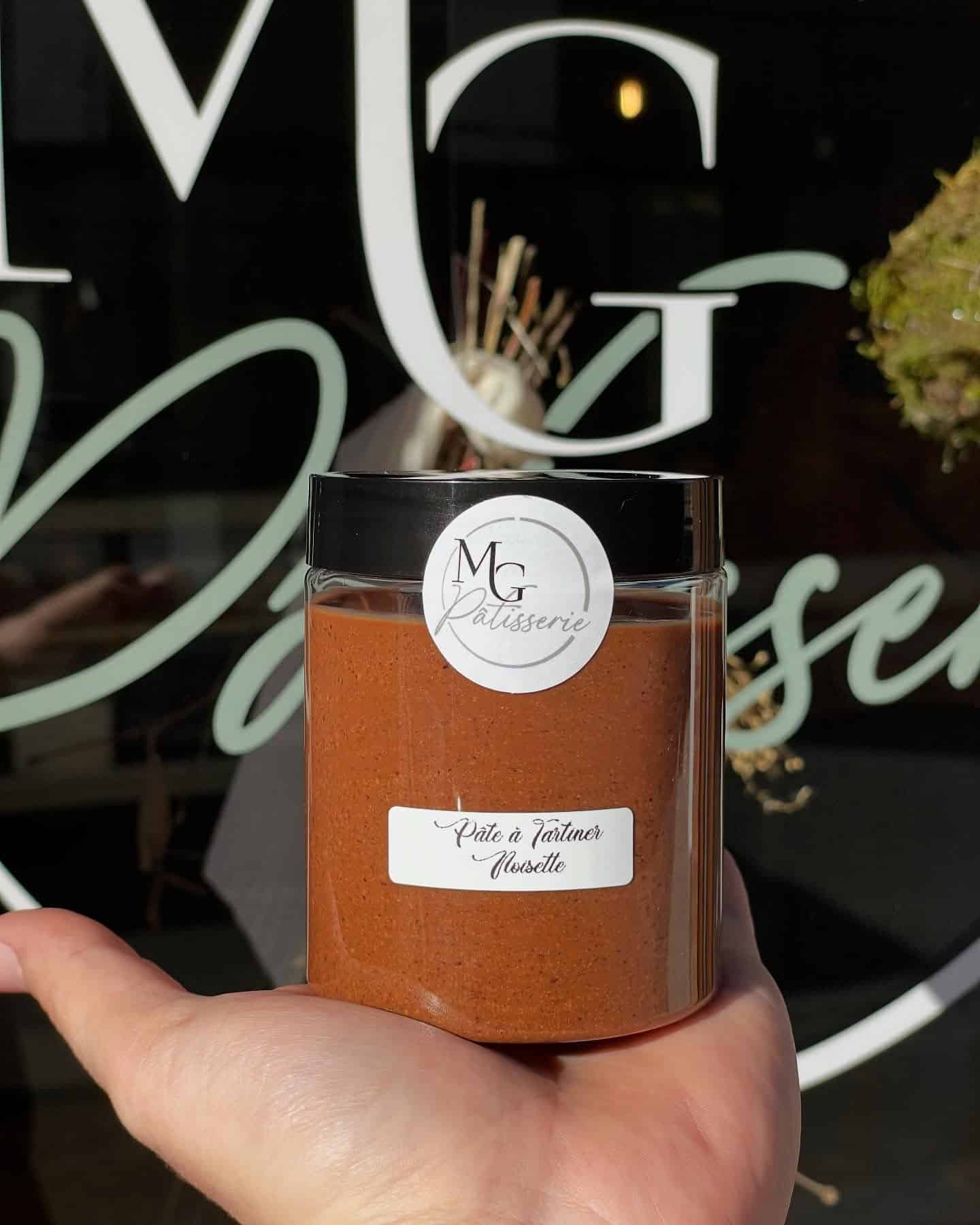 Main tenant un pot de Pâte à tartiner Noisette de MG Pâtisserie, devant une vitrine avec des lettres blanches.