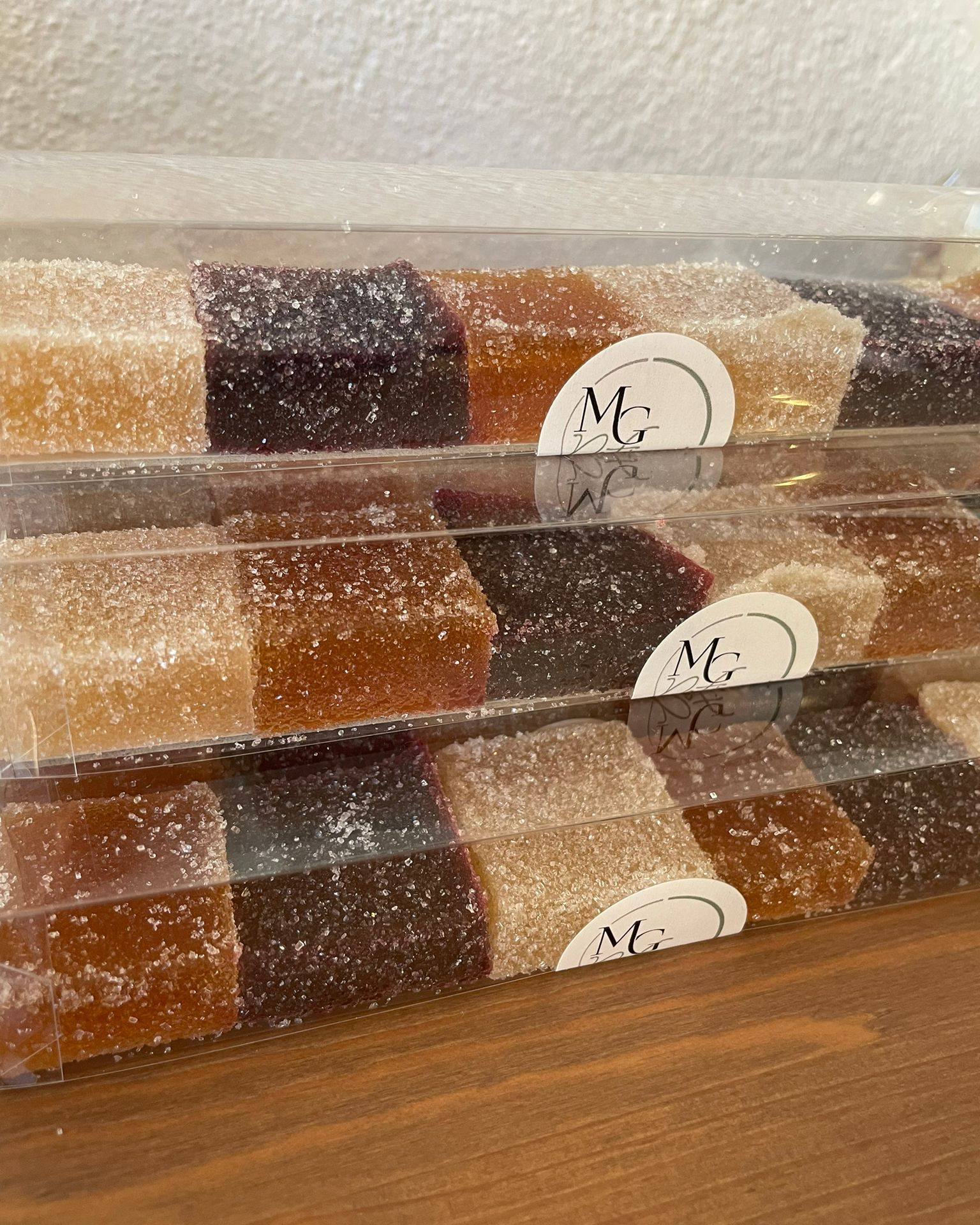 Pâtes de fruits artisanales multicolores, enrobées de sucre, présentées en couches dans une boîte transparente avec un logo 'MG'.