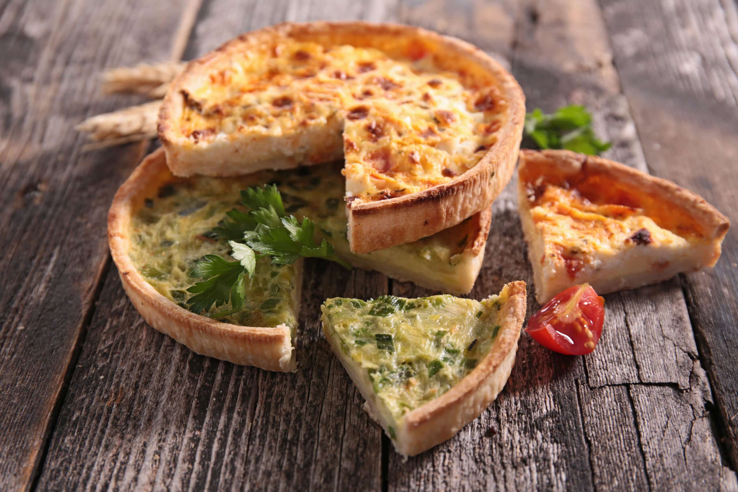Quiches salées variées, dont une aux légumes verts, coupées et présentées sur des planches de bois rustiques avec des brins de blé et une tomate cerise.