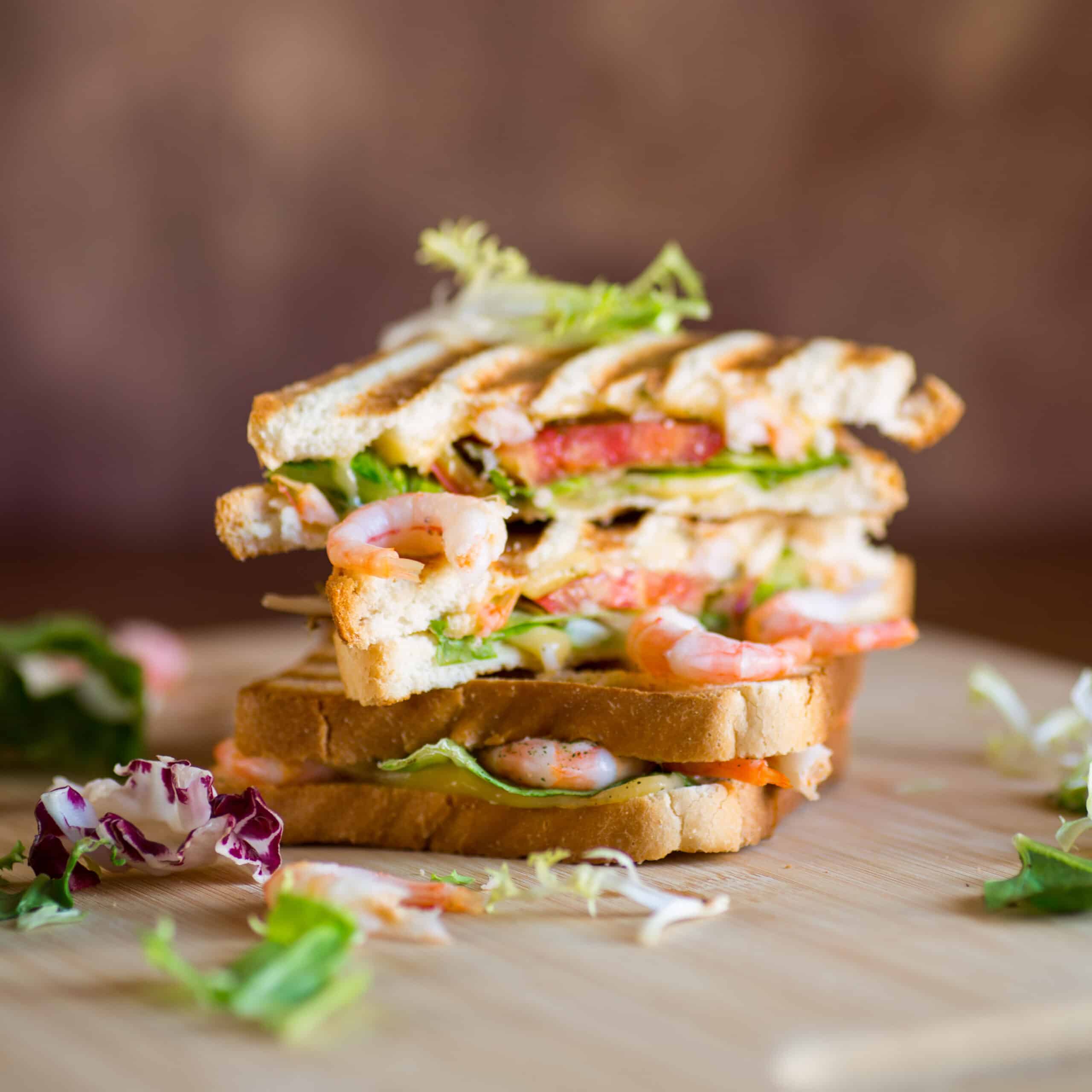 Sandwich club à la crevette grillé, coupé en deux et empilé, garni de laitue et de tomate sur une planche en bois.