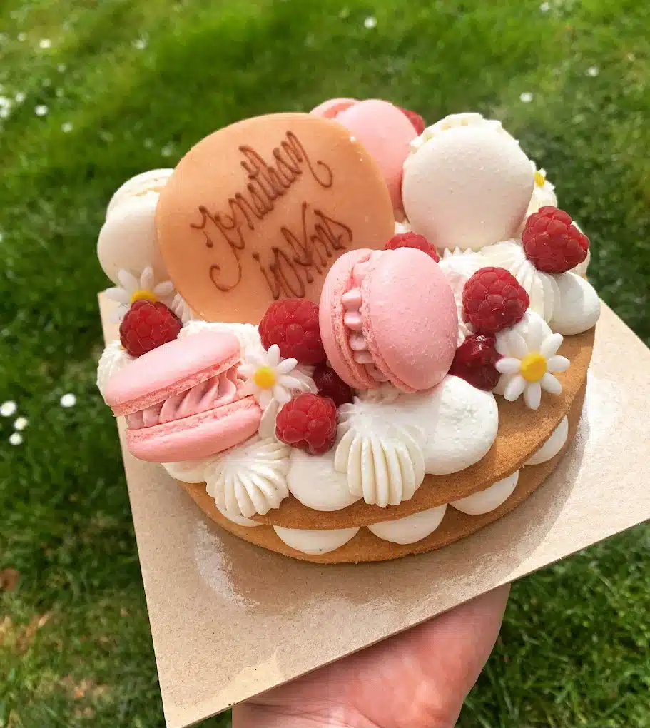 Tartelette biscuit cœur personnalisée 'Jonathan in Wens', décorée de macarons roses, framboises fraîches, crème blanche et petites marguerites, tenue à l'extérieur.