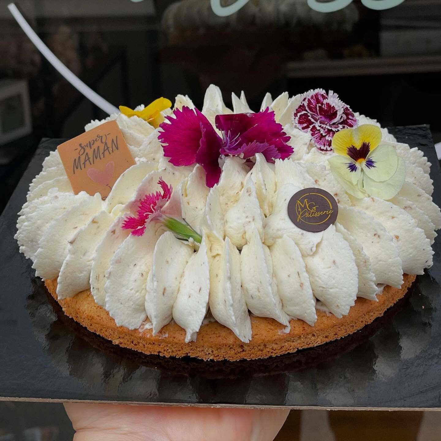 Tartelette gourmande à la crème blanche parsemée de fleurs comestibles et d'une étiquette « Super Maman » tenue à la main.