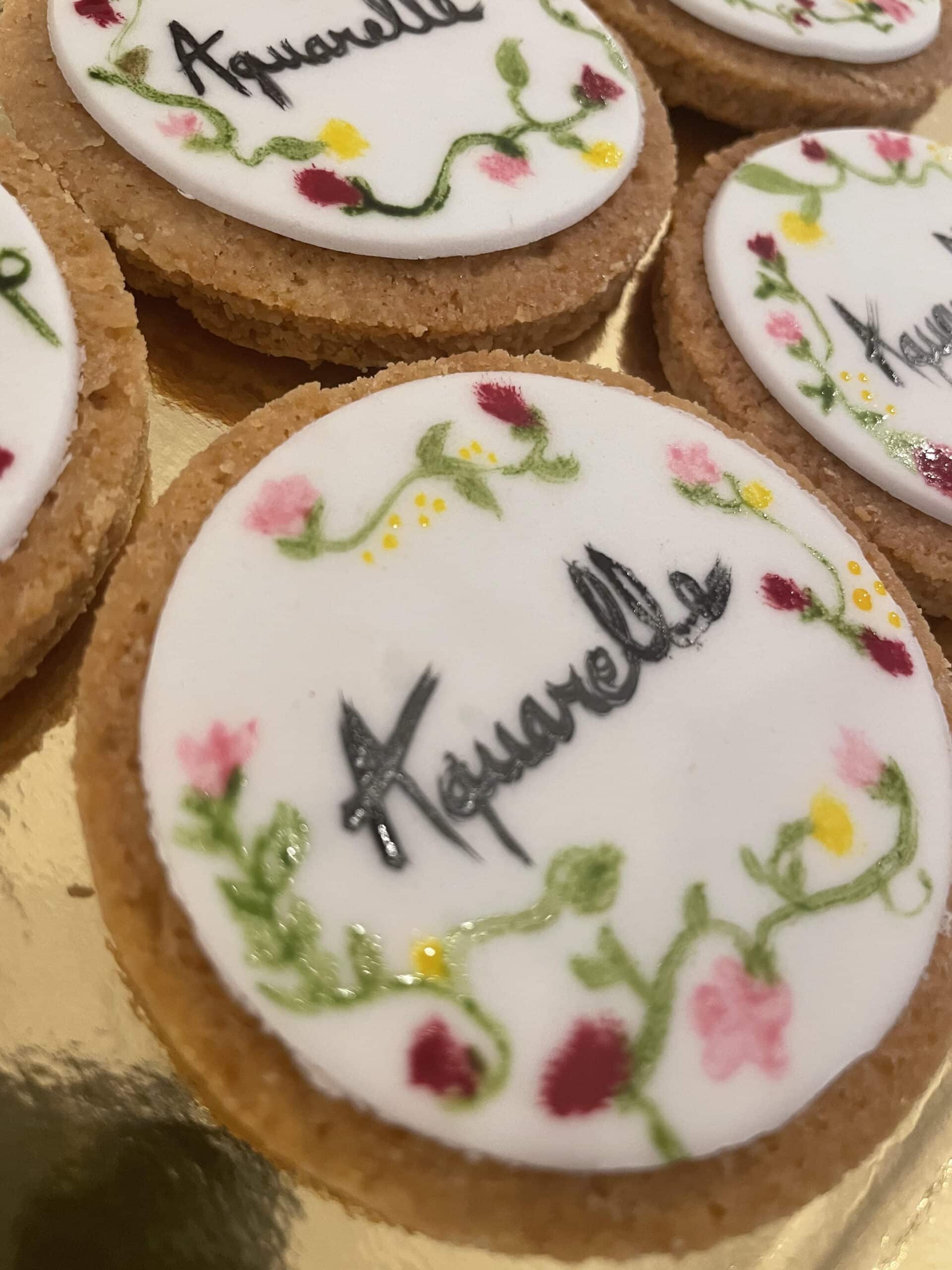 Gros plan sur des biscuits ronds décorés de glaçage blanc. Inscription "Aquarelle" entourée d'une vigne florale peinte rose, rouge et jaune, sur fond doré.