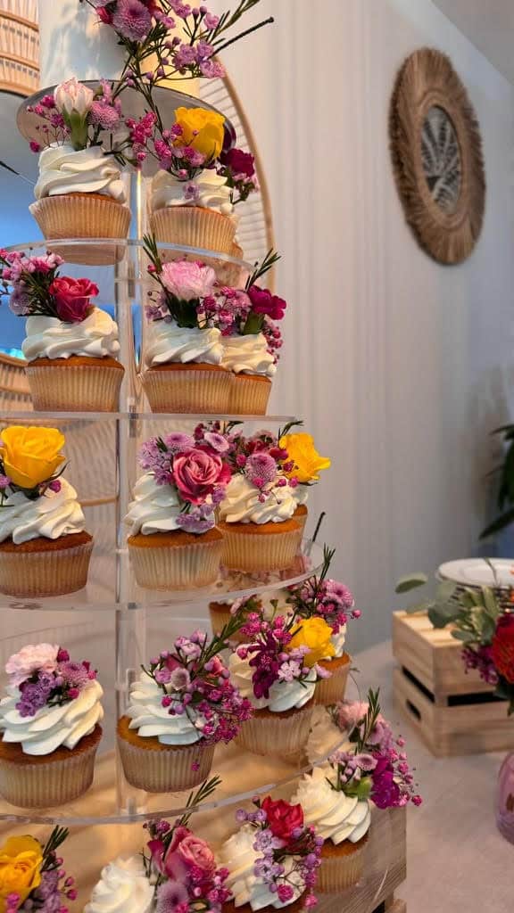 Présentoir à étages de cupcakes à la vanille, nappés de glaçage blanc et ornés de fleurs fraîches variées (roses, jaunes, pourpres).