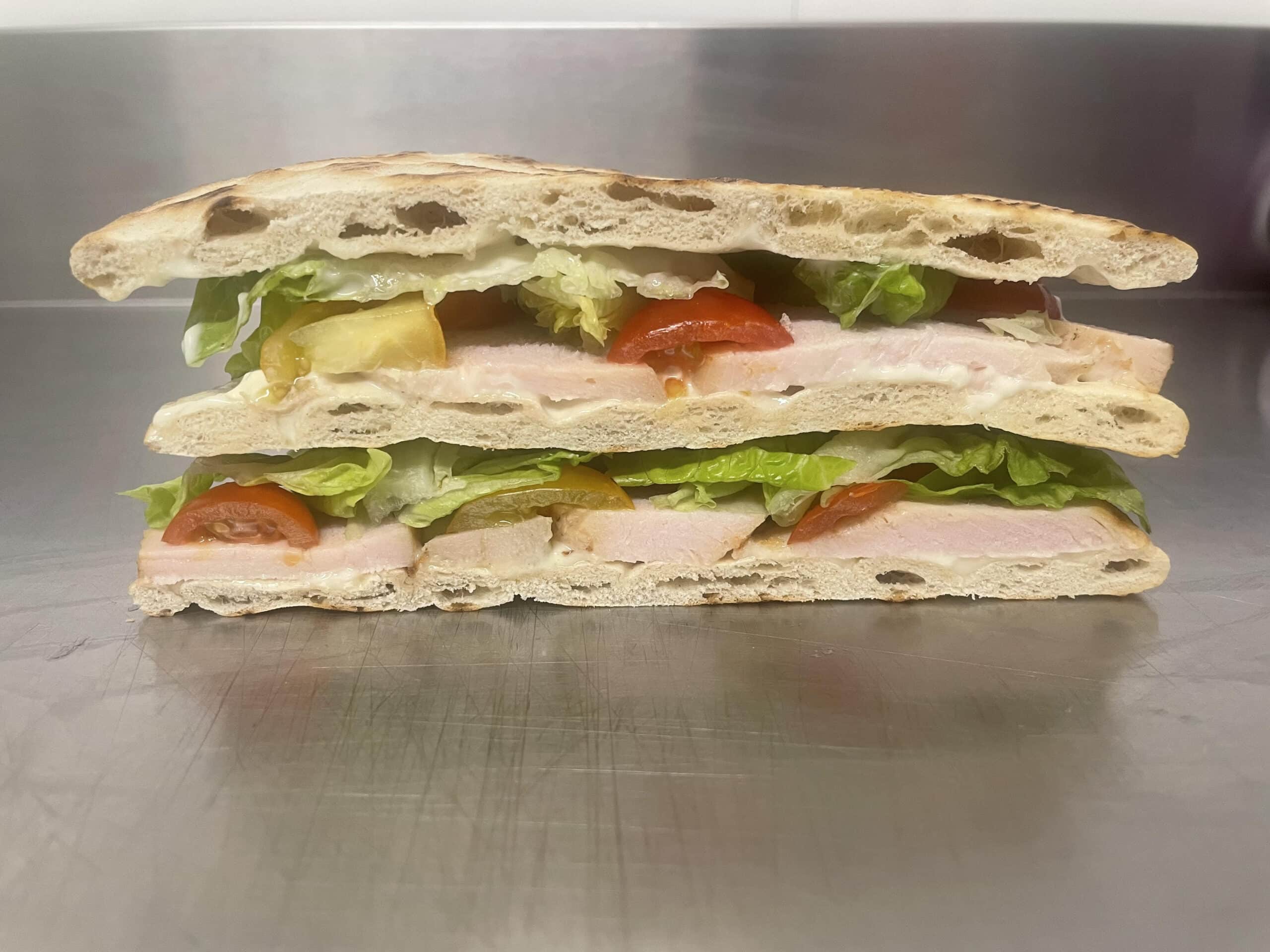 Focaccia toastée, coupée, révélant des couches de dinde ou poulet, laitue fraîche, tomates rouges et jaunes, et sauce blanche.