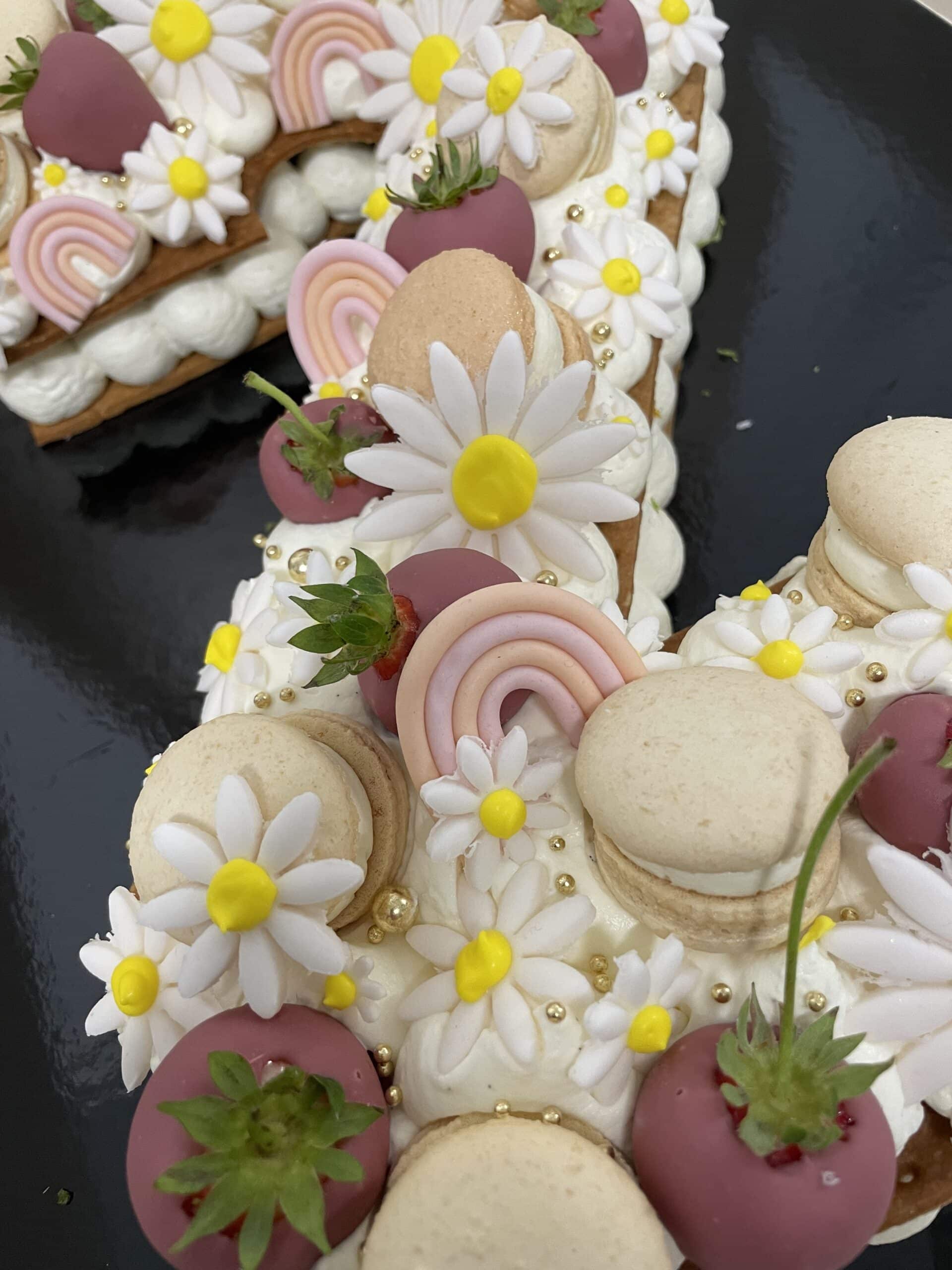 Gâteau tart décoré de macarons, fraises au chocolat mauve, marguerites en fondant, arcs-en-ciel pastel et perles dorées.