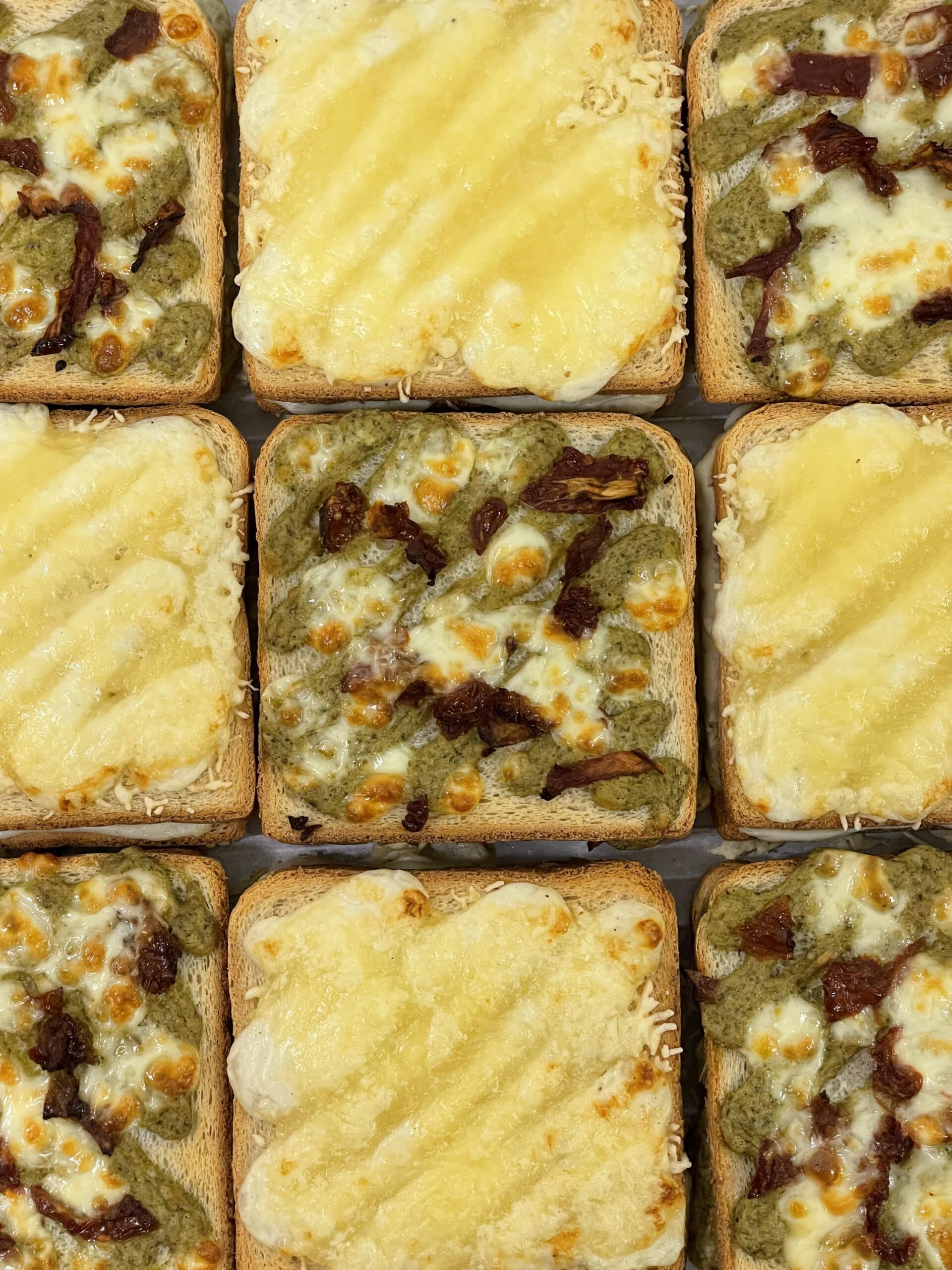 Grille de neuf toasts apéritifs : cinq au pesto, tomates séchées et mozzarella, et quatre entièrement gratinés au fromage fondu.